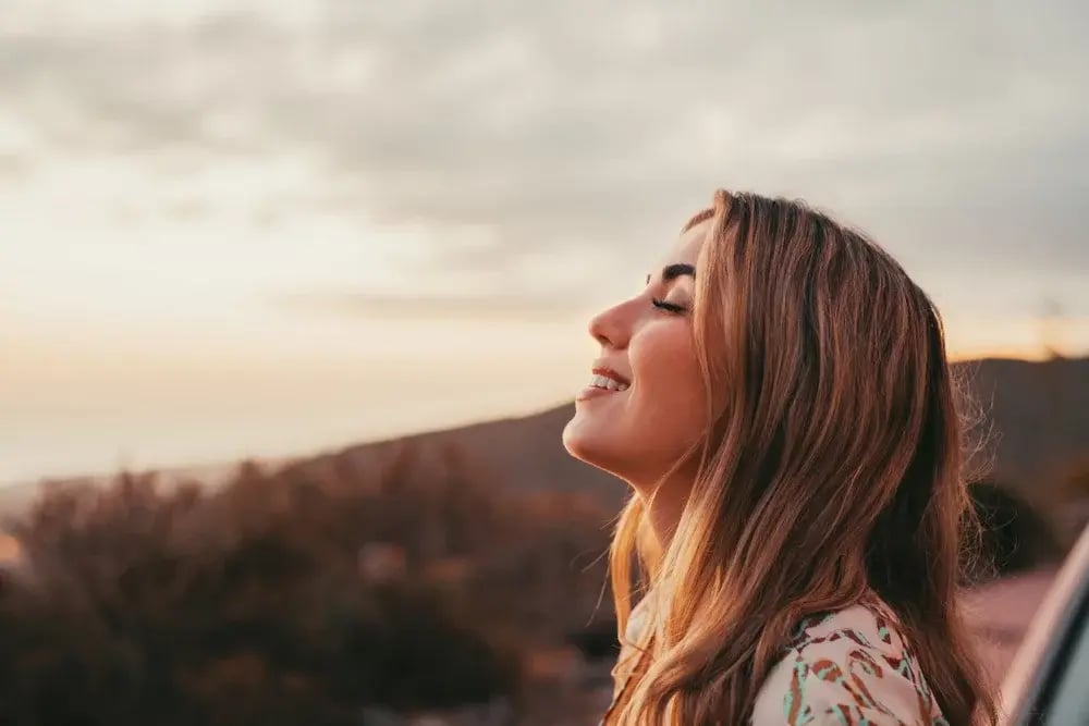 A headshot of a woman standing outside, eyes closed, face toward the sun and smiling.