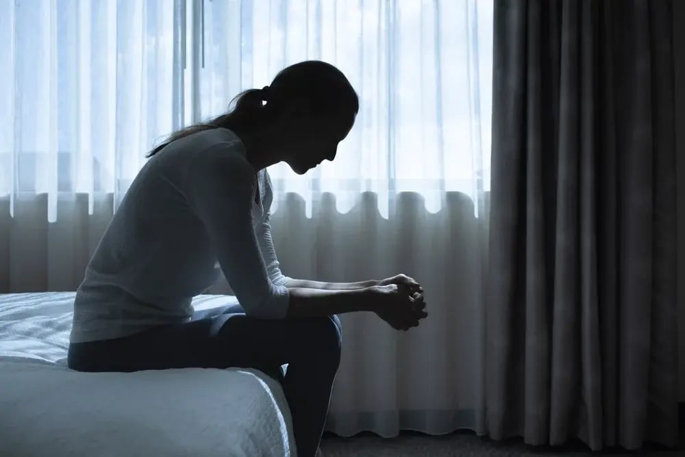 A depressed woman sitting on her bed, staring at her hands.